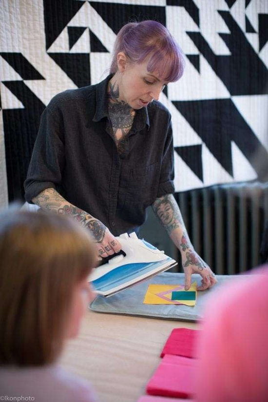 Person with tattoos and purple hair presenting a book or notebook to an audience against a geometric-patterned wall.
