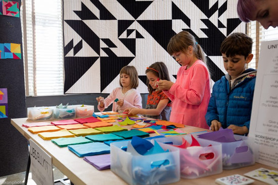 Children engaged in a craft activity at a table with colorful materials, in front of a geometric patterned wall.