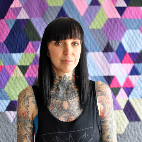 Person with tattoos standing in front of a colorful geometric-patterned quilt
